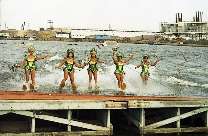 Historic photo from Saturday, August 29, 1970 - 5 women waterskiing towards jump, holding flowers, with Ontario Place bridge and pavilion pods under construction in background - CNE Aquarama show in Ontario Place