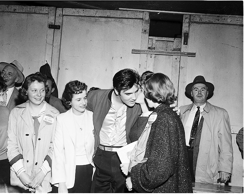 Elvis standing with group of female fans; some of the fans are wearing badges, Elvis has tilted his head to get a kiss on the cheek from one of the fans. Image no. ASC00846.